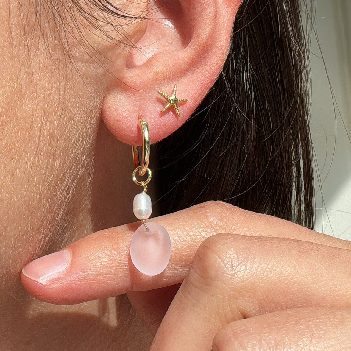 Close-up of an ear with a gold earring featuring a star and pink gemstone, held by a hand.