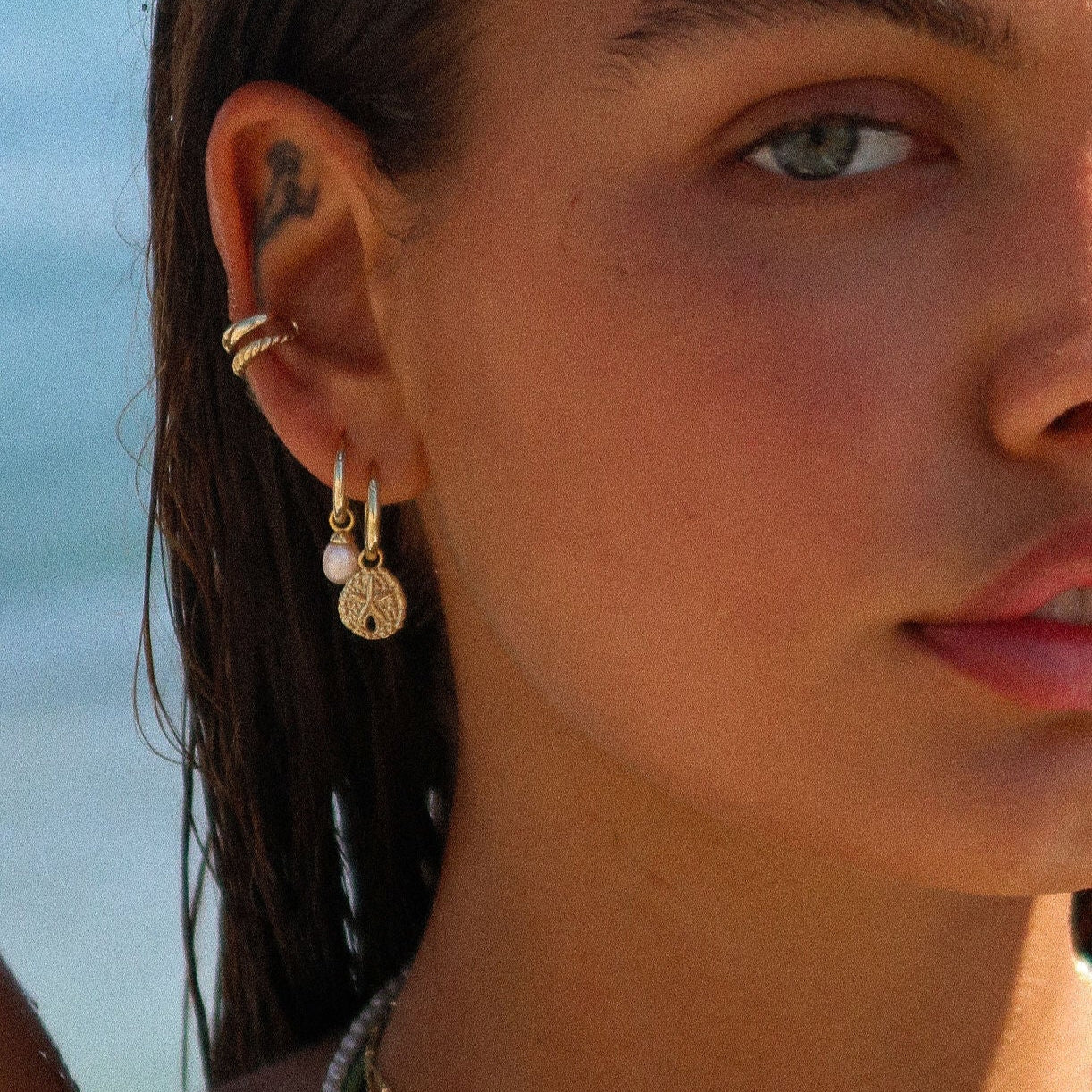 Close-up of a woman's ear with gold earrings and rings, against a blurred natural background.
